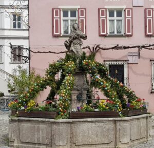 Ein historischer Brunnen mit Marienstatue, festlich mit bunten Ostereiern geschmückt.