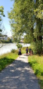 Eine Frau im dunklen Kleid geht entspannt den Kiesweg am Fluss entlang.