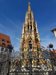 Der goldverzierte gotische Brunnenturm ragt eindrucksvoll in den blauen Himmel.
