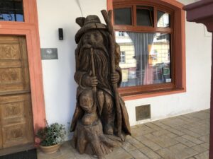 Geschnitzte Holzfigur mit Hut und Wanderstab vor einem Hauseingang